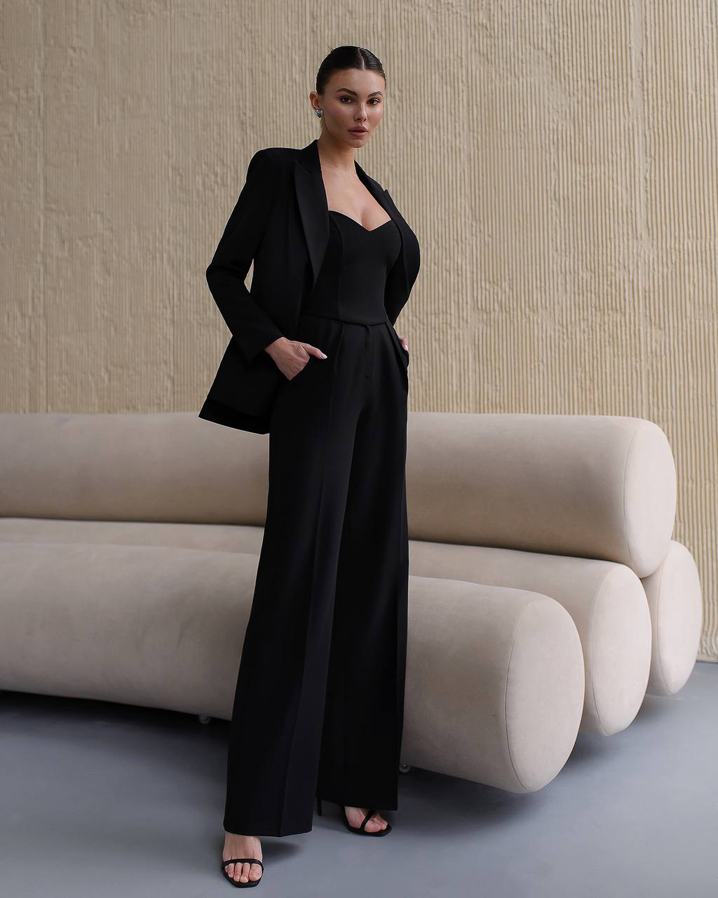 Woman in a black suit standing in front of a beige textured wall with a modern sofa.