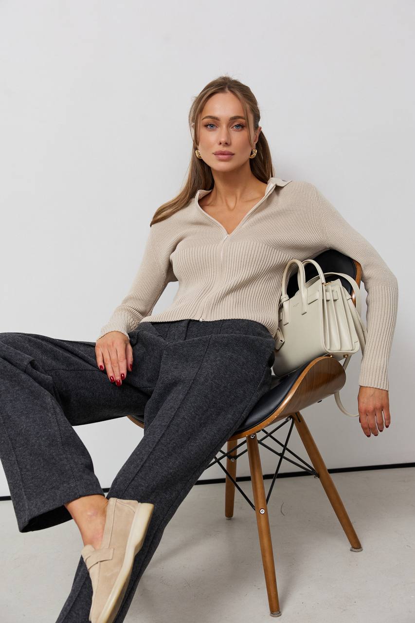 Woman sitting on a chair wearing a beige sweater and dark pants, with a white background