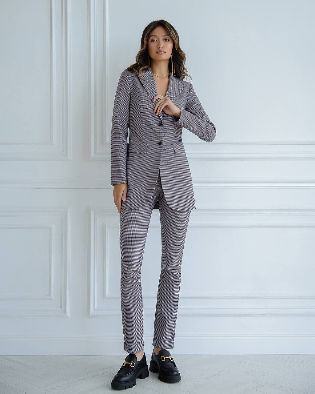Woman wearing a gray suit with black shoes against a white paneled wall.
