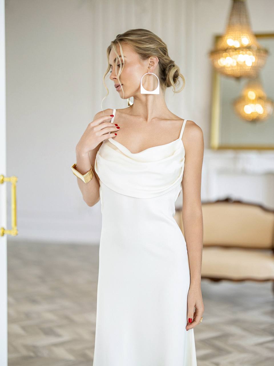 A woman wearing a white cowl-neck spaghetti strap dress, styled with large white hoop earrings and a gold bracelet, standing in a brightly lit, elegant room.