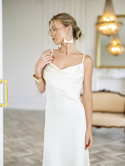 A woman wearing a white cowl-neck spaghetti strap dress, styled with large white hoop earrings and a gold bracelet, standing in a brightly lit, elegant room.