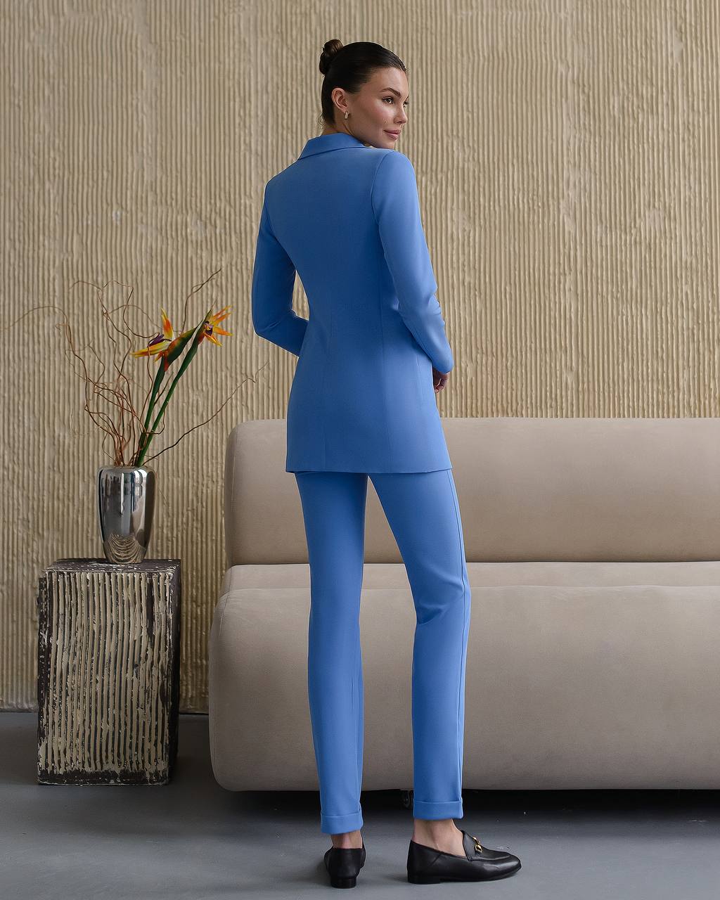 Woman in a blue suit standing in a room with a beige sofa and decorative plant.