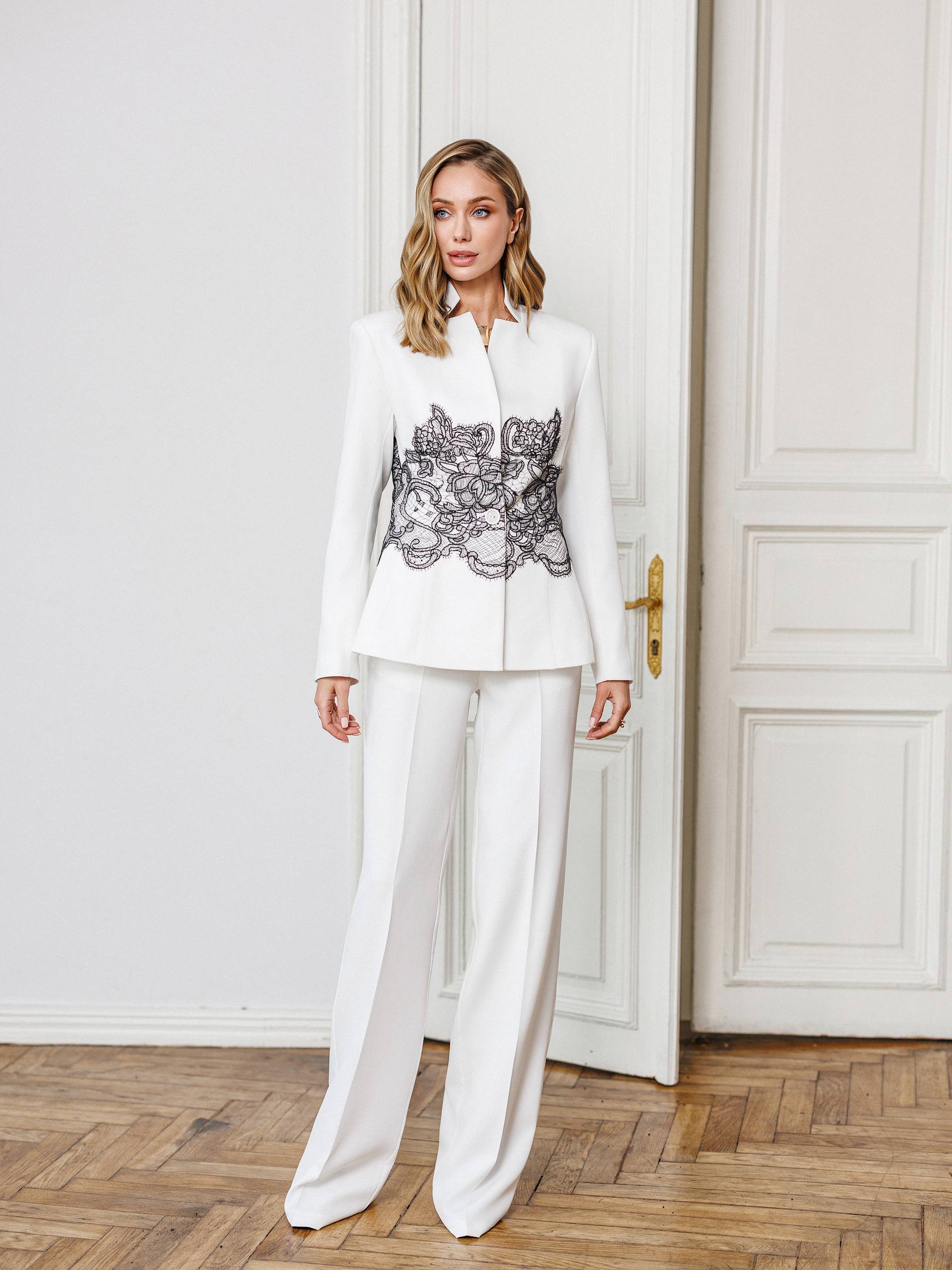 Woman wearing a white suit with an embroidered top in a room with wooden flooring and white walls.