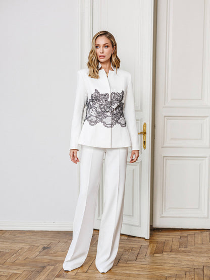 Woman wearing a white suit with an embroidered top in a room with wooden flooring and white walls.