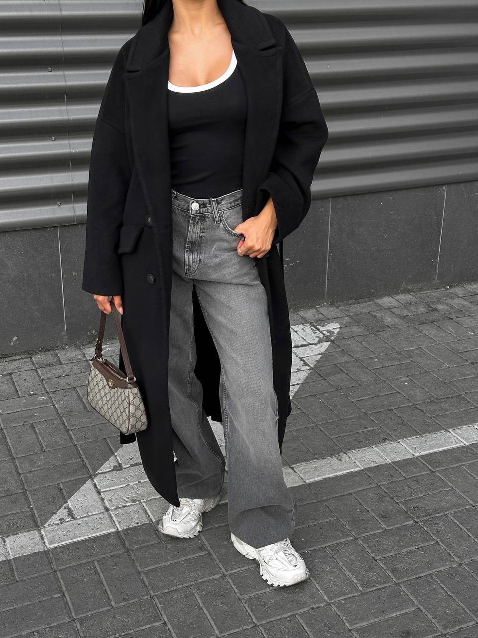 Person wearing a black coat, black top, gray jeans, and white sneakers on a sidewalk.