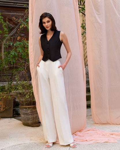 Woman wearing a black sleeveless top and white pants standing in front of pink curtains.