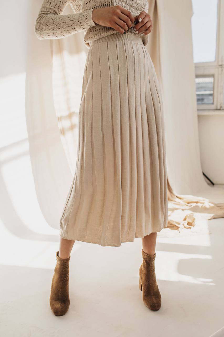 Person wearing a beige pleated skirt and brown ankle boots in a bright room.
