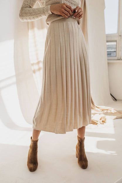 Person wearing a beige pleated skirt and brown ankle boots in a bright room.