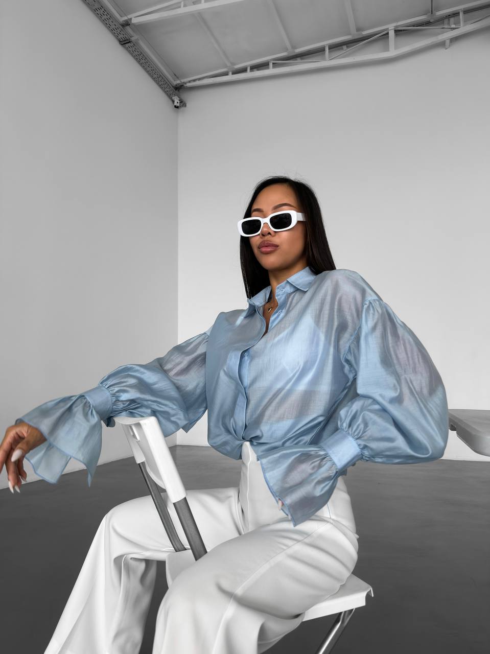 Person wearing a light blue shirt and white pants sitting on a chair in a minimalistic setting.
