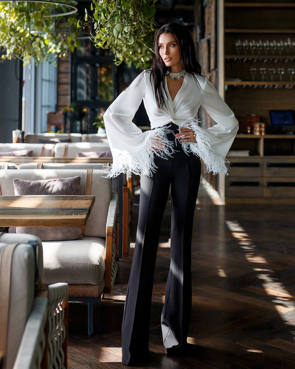 Woman in a white blouse with fringe details and black pants standing in a restaurant.