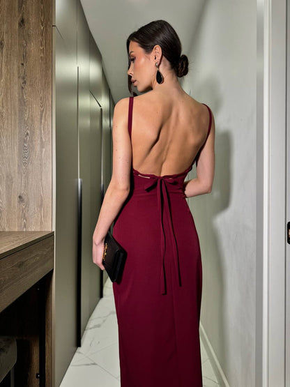 Woman in a burgundy evening gown with an open back, standing in a hallway.