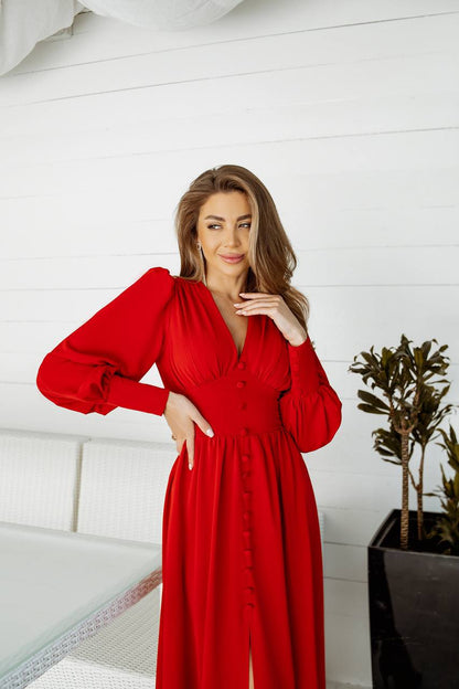Woman wearing a red dress standing indoors with a white wall and plant in the background