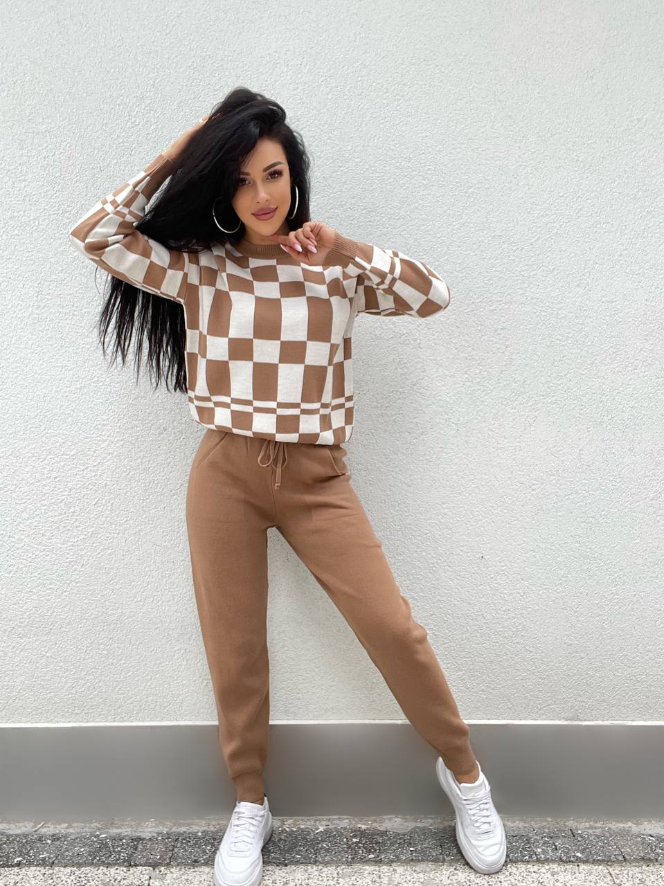 Woman wearing a checkered sweater and brown pants against a light gray wall.