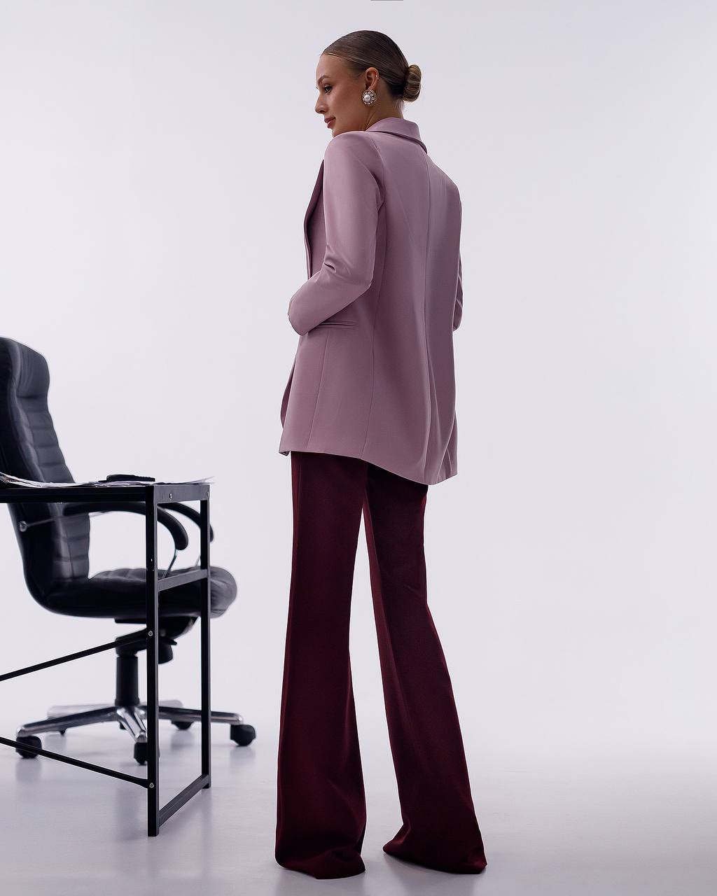 A woman stands in profile wearing a lavender blazer and dark burgundy wide-leg trousers in a minimalist studio setting with a black office chair on the left.