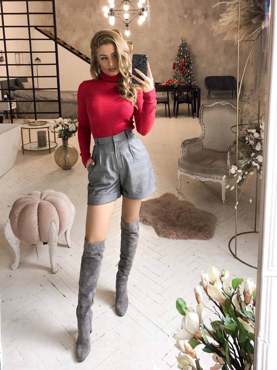 Person taking a mirror selfie wearing a red turtleneck, gray shorts, and gray knee-high boots in a stylish room.