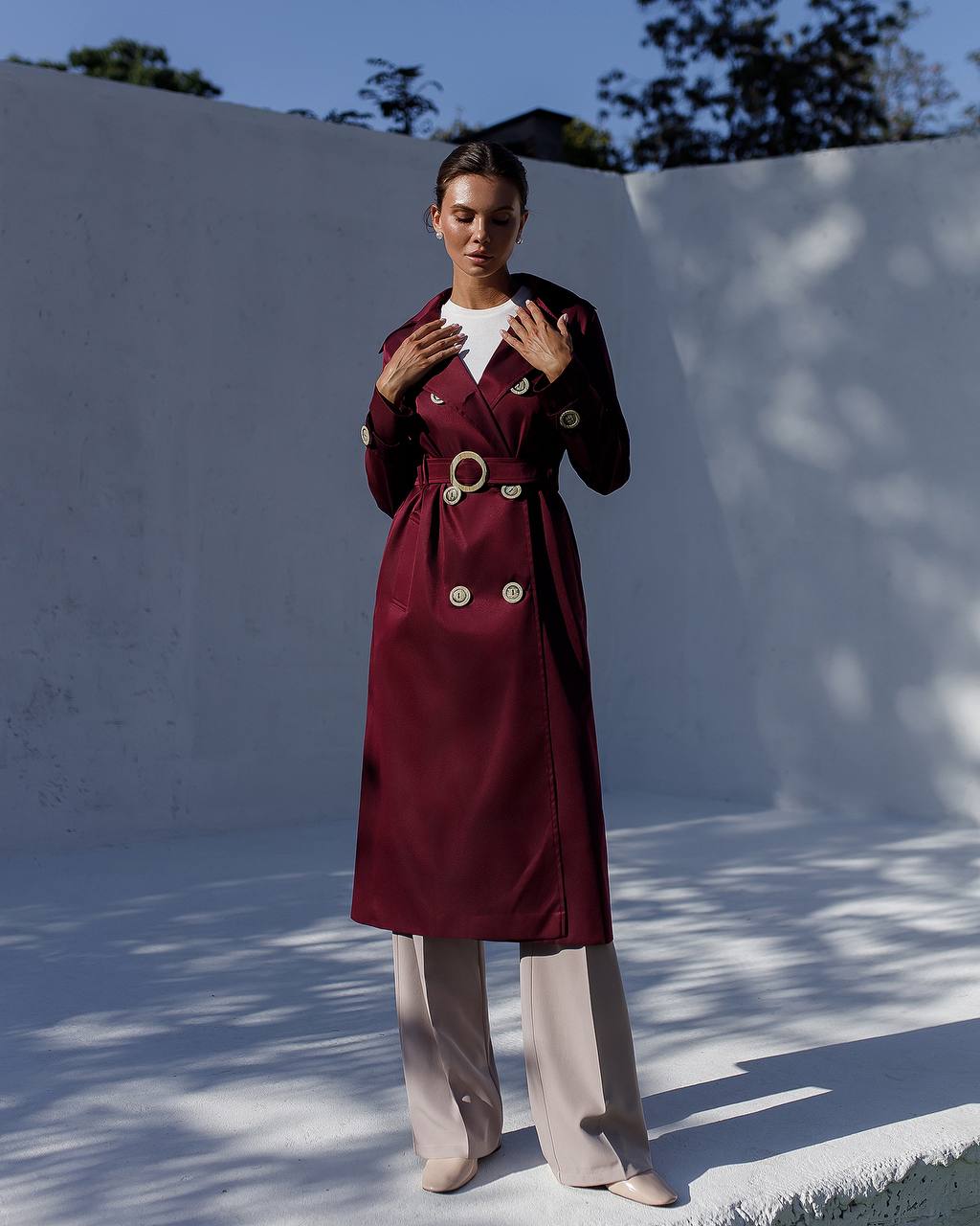 Person wearing a burgundy trench coat with gold buttons against a white wall.