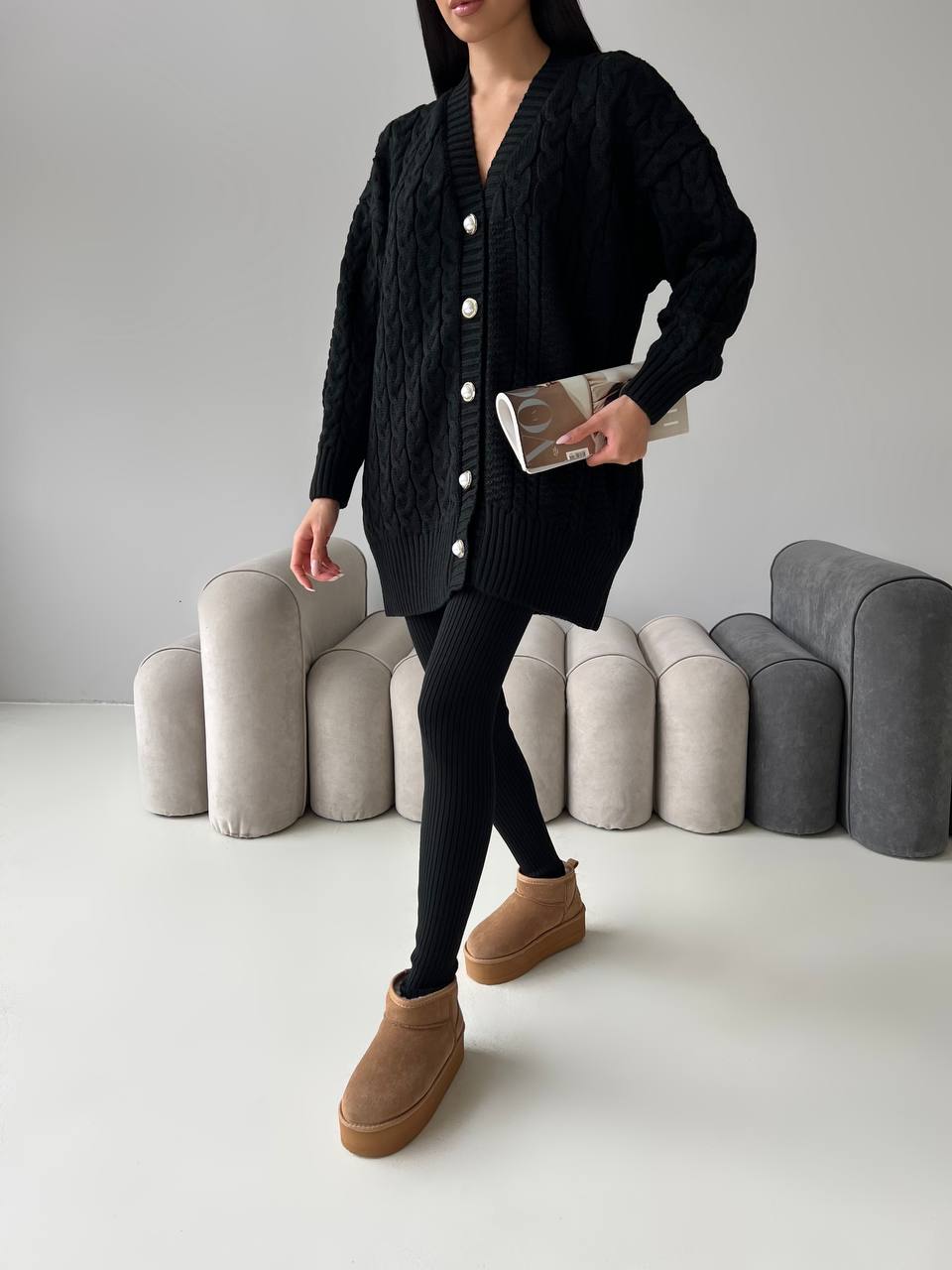Person wearing a black cardigan, black leggings, and brown boots in a minimalistic setting with gray cushions.