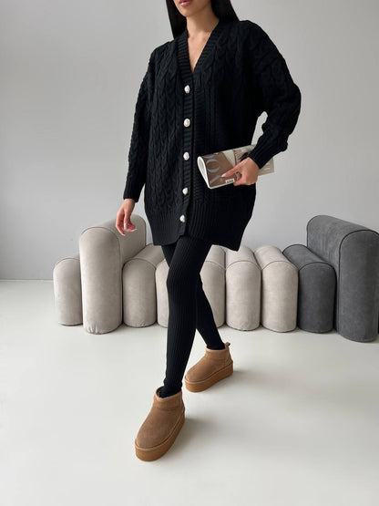 Person wearing a black cardigan, black leggings, and brown boots in a minimalistic setting with gray cushions.
