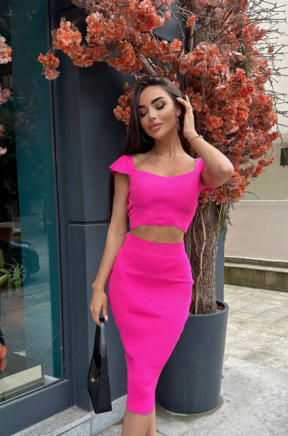 Woman in a bright pink outfit standing outdoors next to a tree with orange flowers.