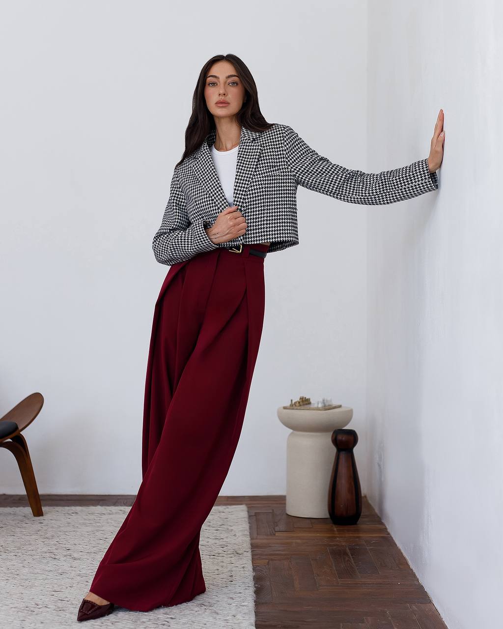 Woman wearing a patterned jacket and burgundy pants in a minimalistic room.