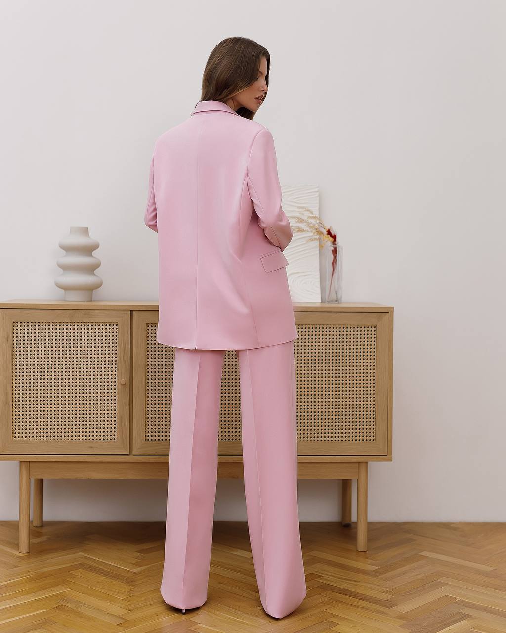 Woman in a pink suit standing in a room with wooden floor and light-colored wall.
