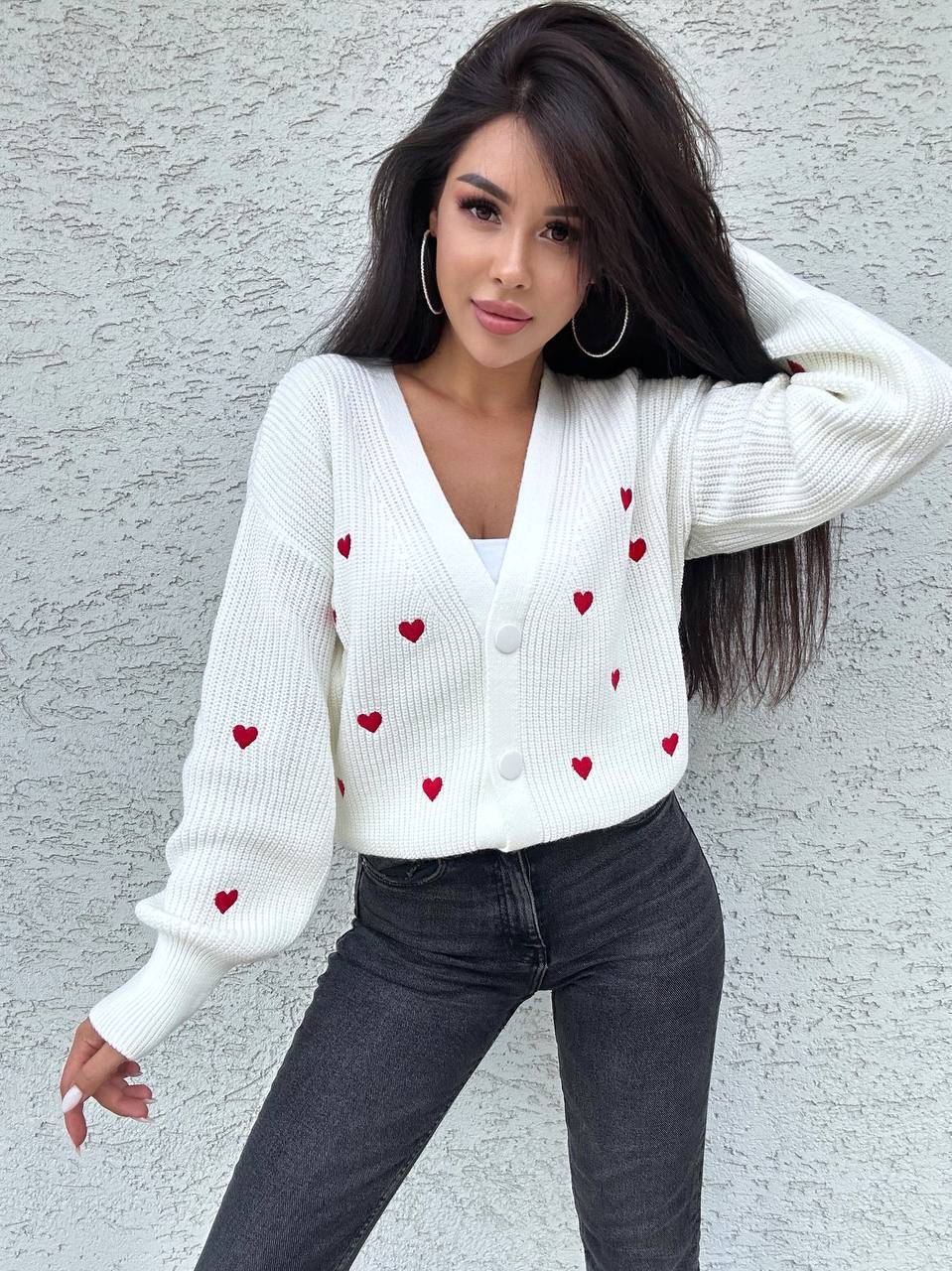 Woman wearing a white sweater with red heart patterns against a gray wall.
