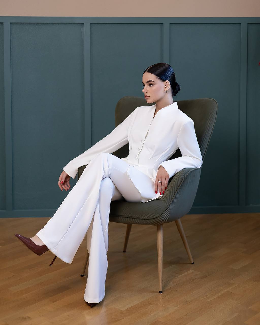 Woman in a white outfit sitting on a modern chair against a teal wall.