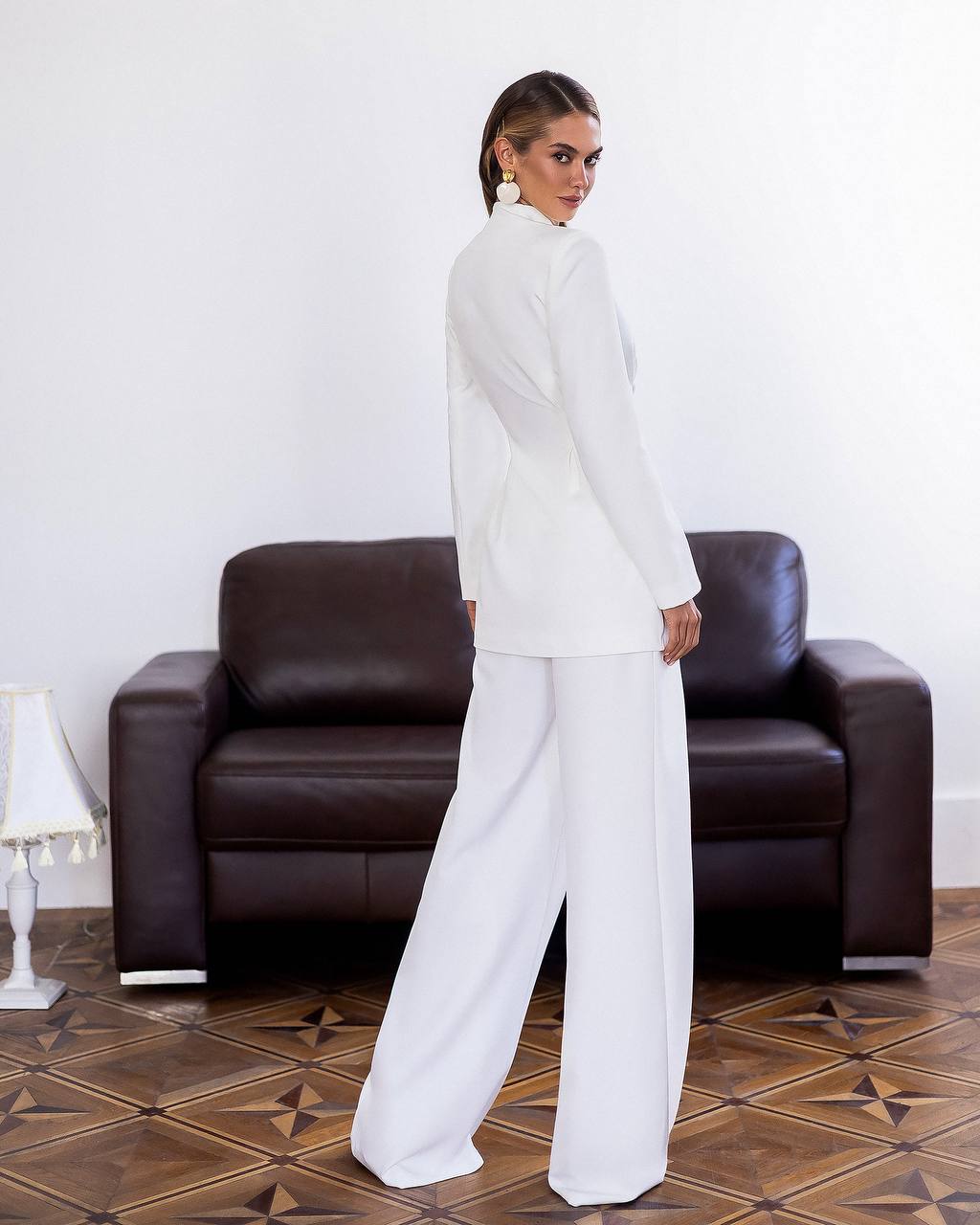 Woman in a white outfit standing in front of a brown leather armchair.