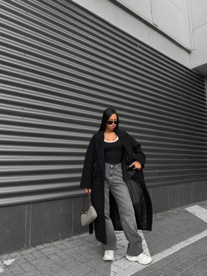 Person wearing a long black coat, black top, gray pants, and white sneakers against a corrugated metal wall.
