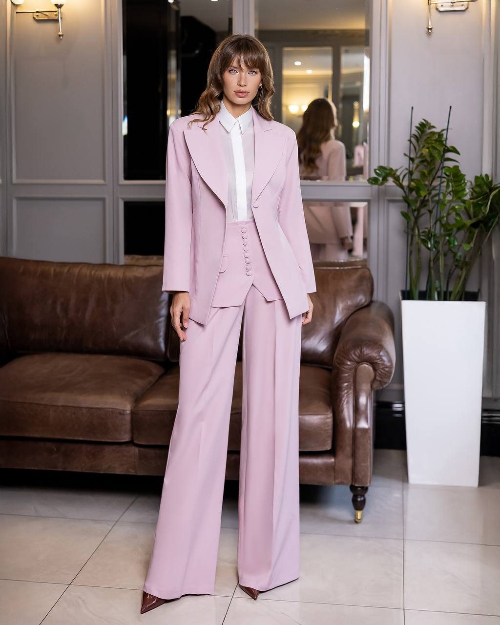 Woman in a pink suit standing in a room with a brown leather couch and a plant.