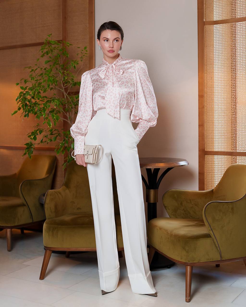 Woman in a stylish outfit standing in an elegant room with green chairs and a plant.