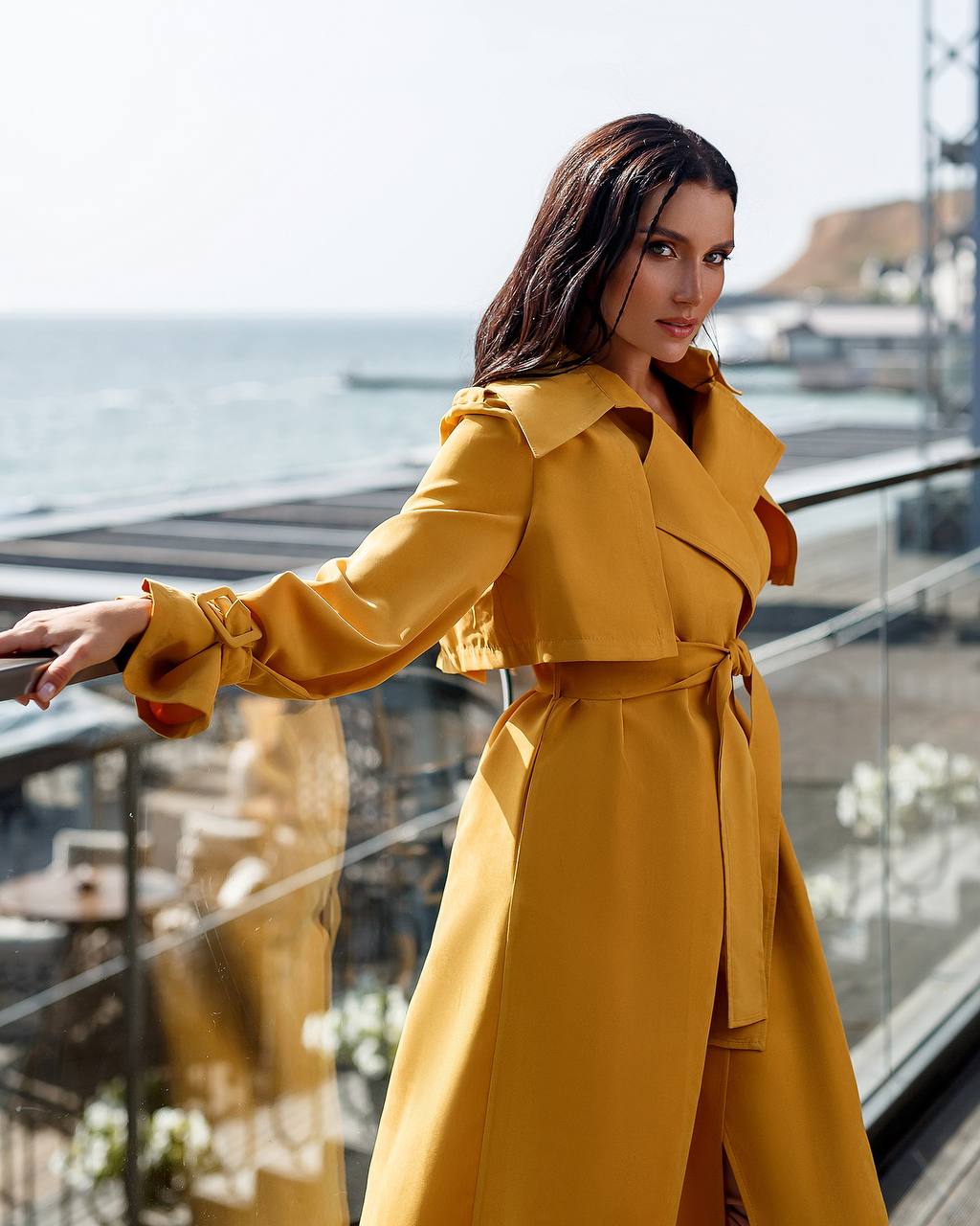 Woman wearing a yellow trench coat standing by a waterfront.