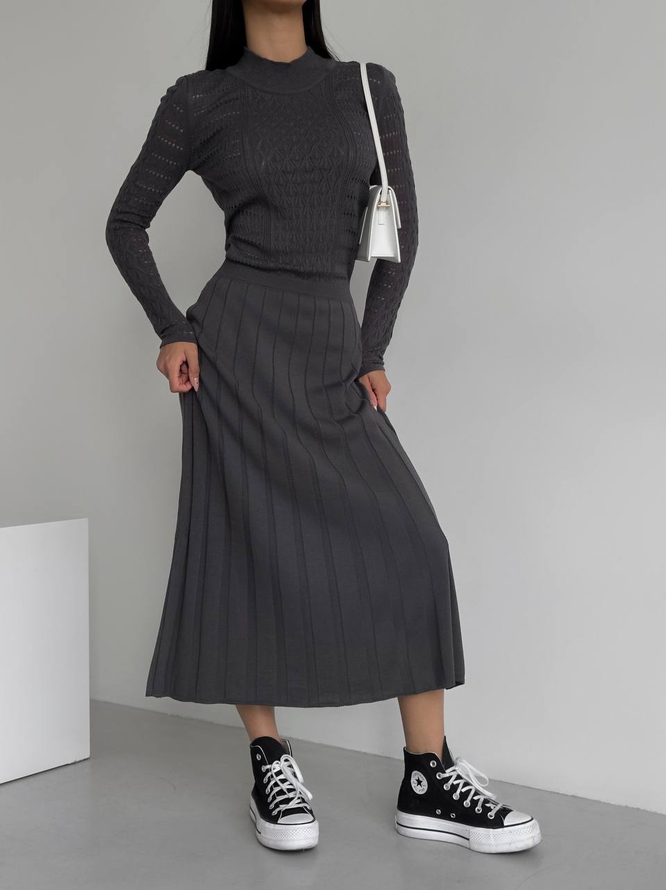 Person wearing a dark gray ribbed knit outfit with a white bag on a plain background