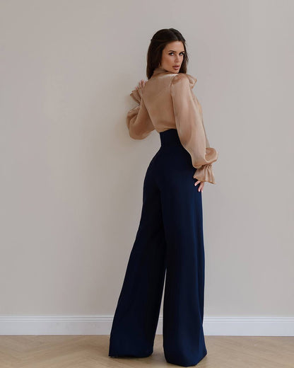 Woman wearing a beige blouse and navy wide-leg pants against a plain wall.