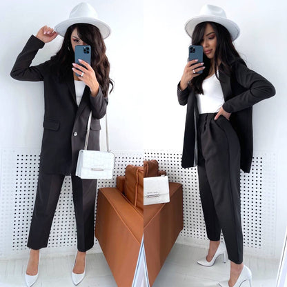 Woman in a black suit and white hat taking a mirror selfie in a minimalistic setting.