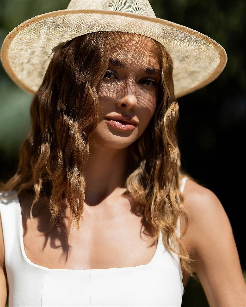A woman wearing a wide-brimmed straw hat with a sheer black mesh sun veil and a white sleeveless top, photographed outdoors in bright sunlight against a dark green blurred background.