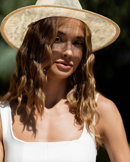 A woman wearing a wide-brimmed straw hat with a sheer black mesh sun veil and a white sleeveless top, photographed outdoors in bright sunlight against a dark green blurred background.