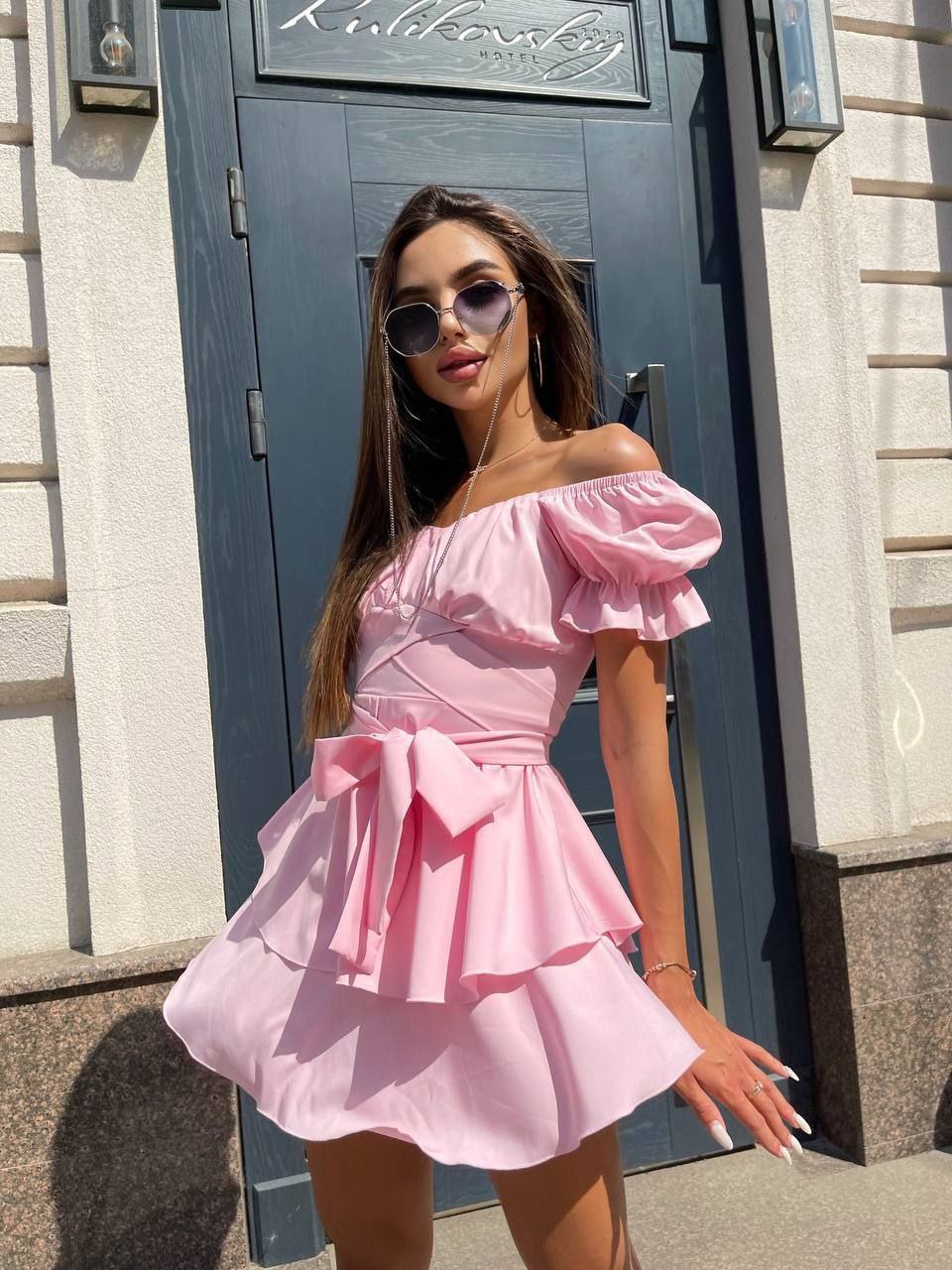 Woman wearing a pink off-shoulder dress standing in front of a building entrance.