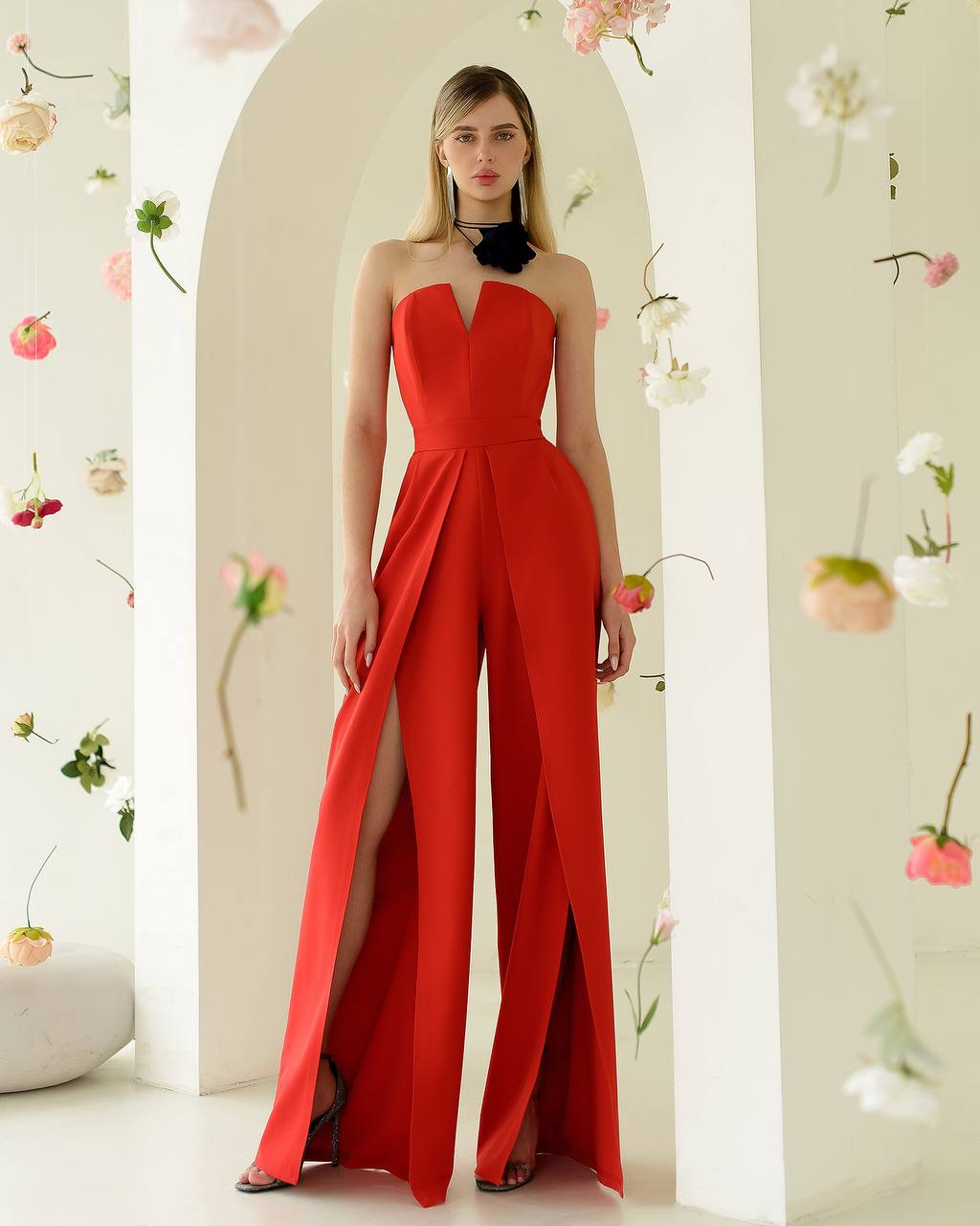 Woman wearing a strapless red jumpsuit with a high slit, standing against a floral decorated wall.