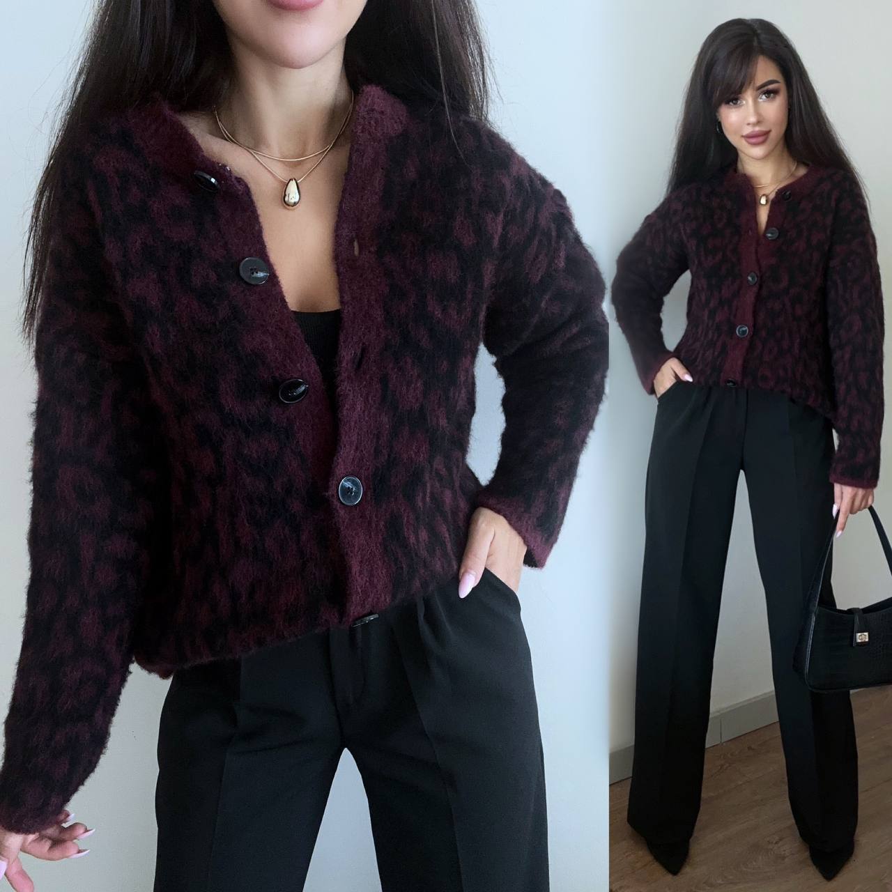 Woman wearing a patterned cardigan and black pants against a white wall.