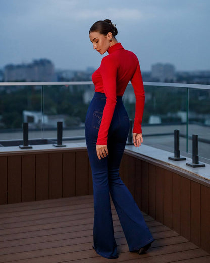Woman in red top and blue flared pants standing on a rooftop with cityscape view.