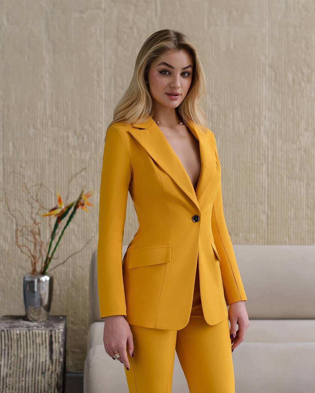 Woman wearing a mustard yellow suit standing in an indoor setting with a neutral background.