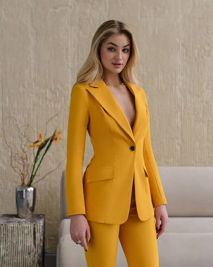 Woman wearing a mustard yellow suit standing in an indoor setting with a neutral background.