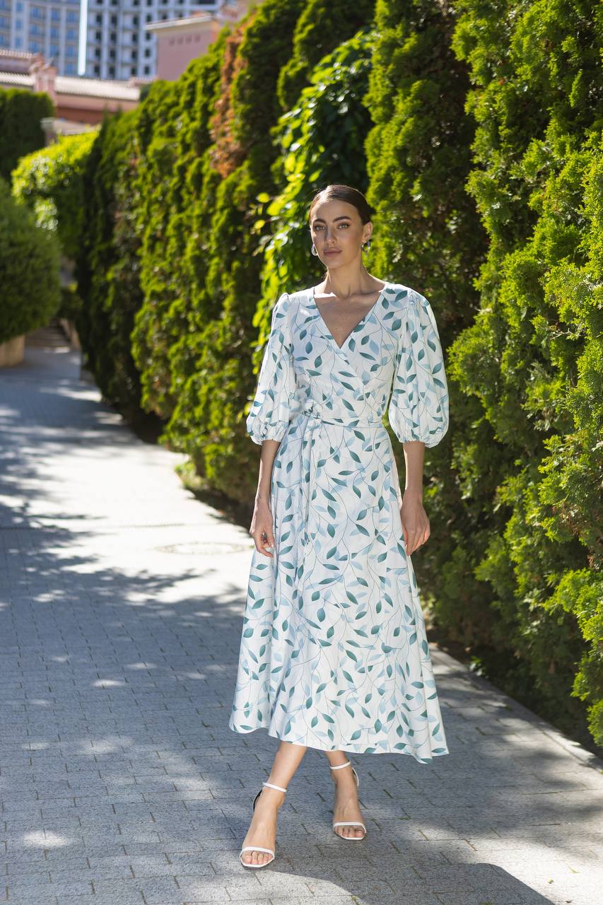 Woman in a floral dress standing on a sidewalk with greenery in the background