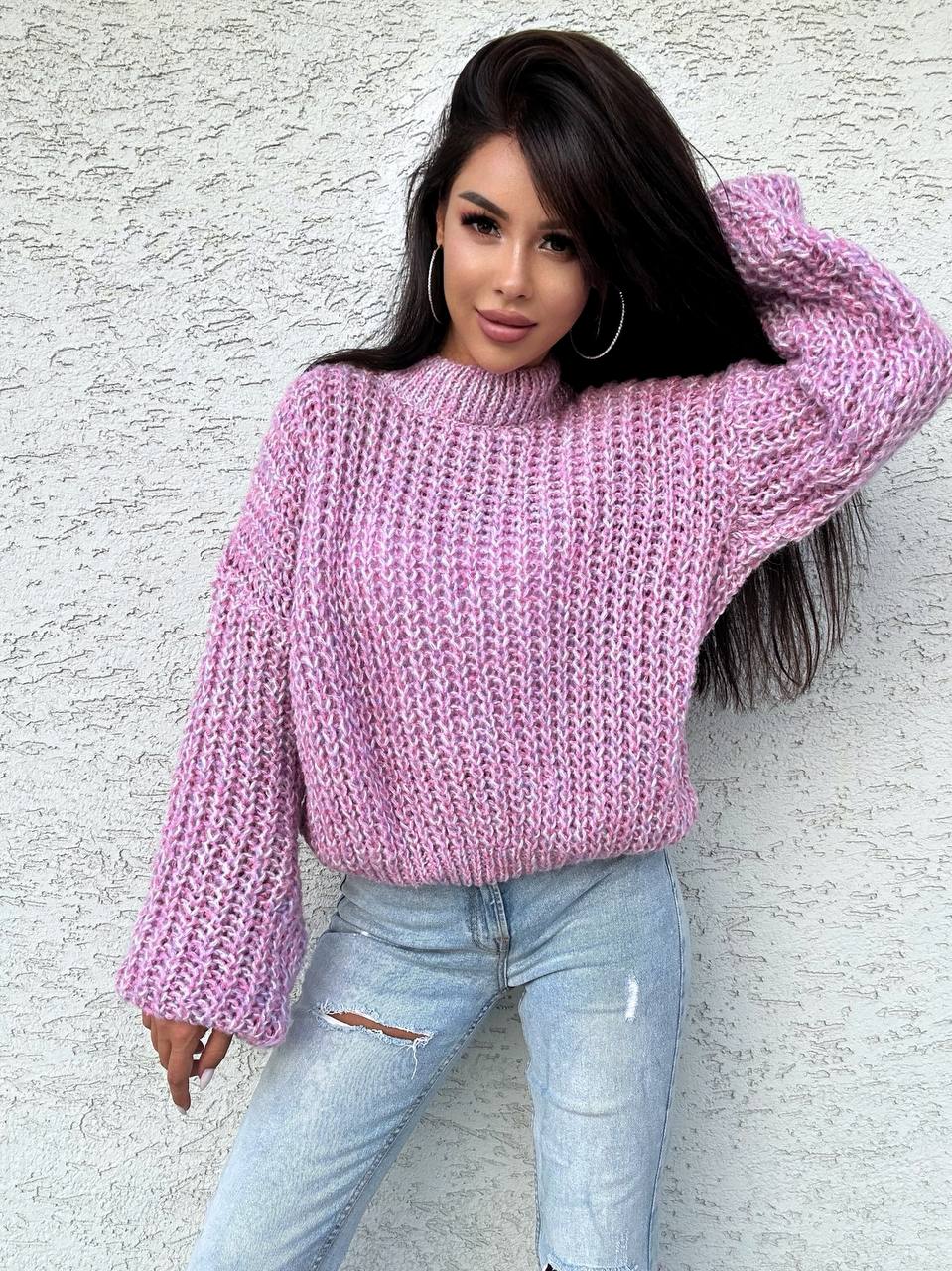 Woman wearing a pink knitted sweater and light blue jeans against a white wall.