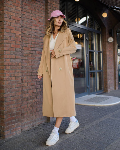 Woman wearing a beige coat and pink hat standing against a brick wall.
