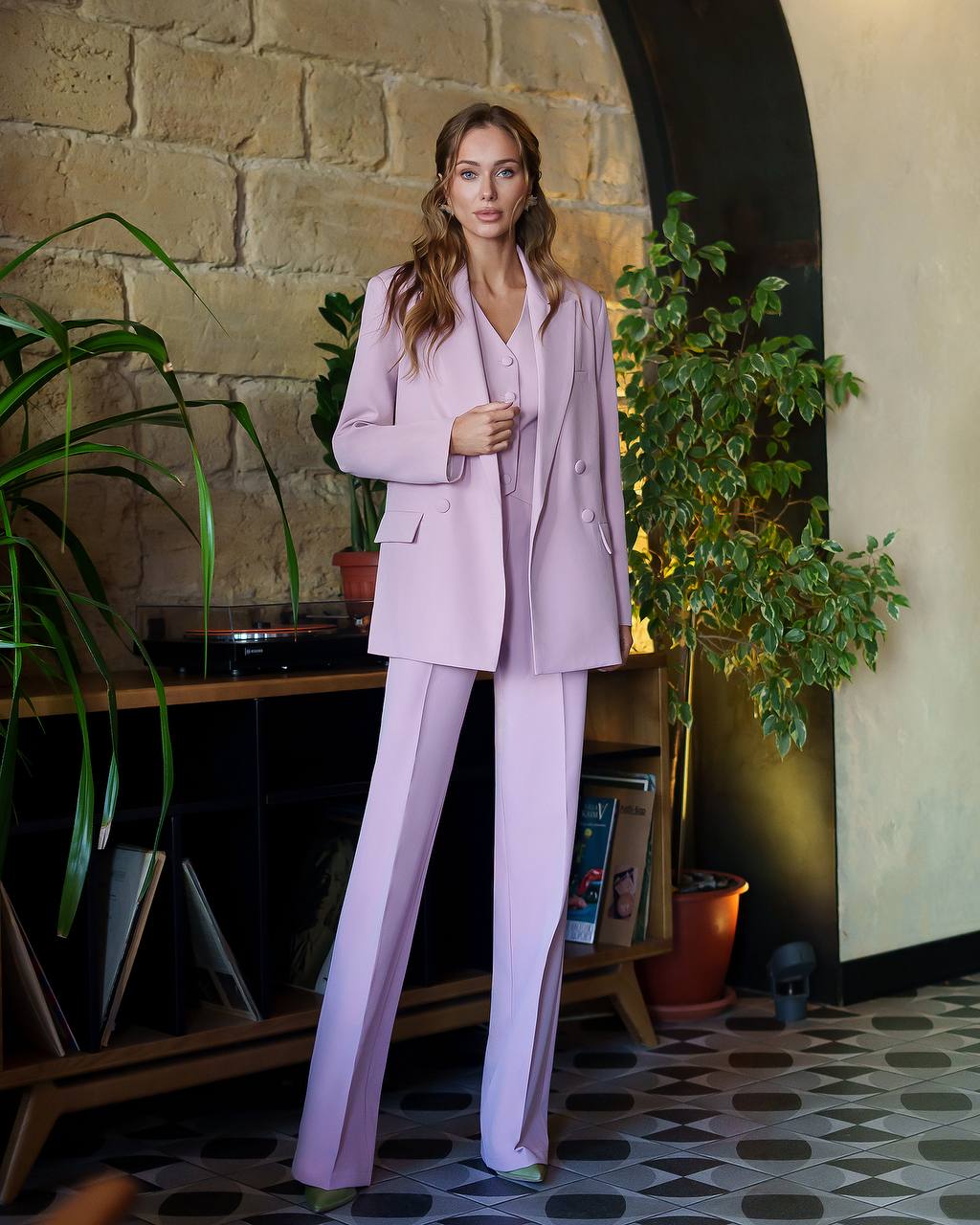 Woman in a lavender suit standing in a room with plants and a stone wall.