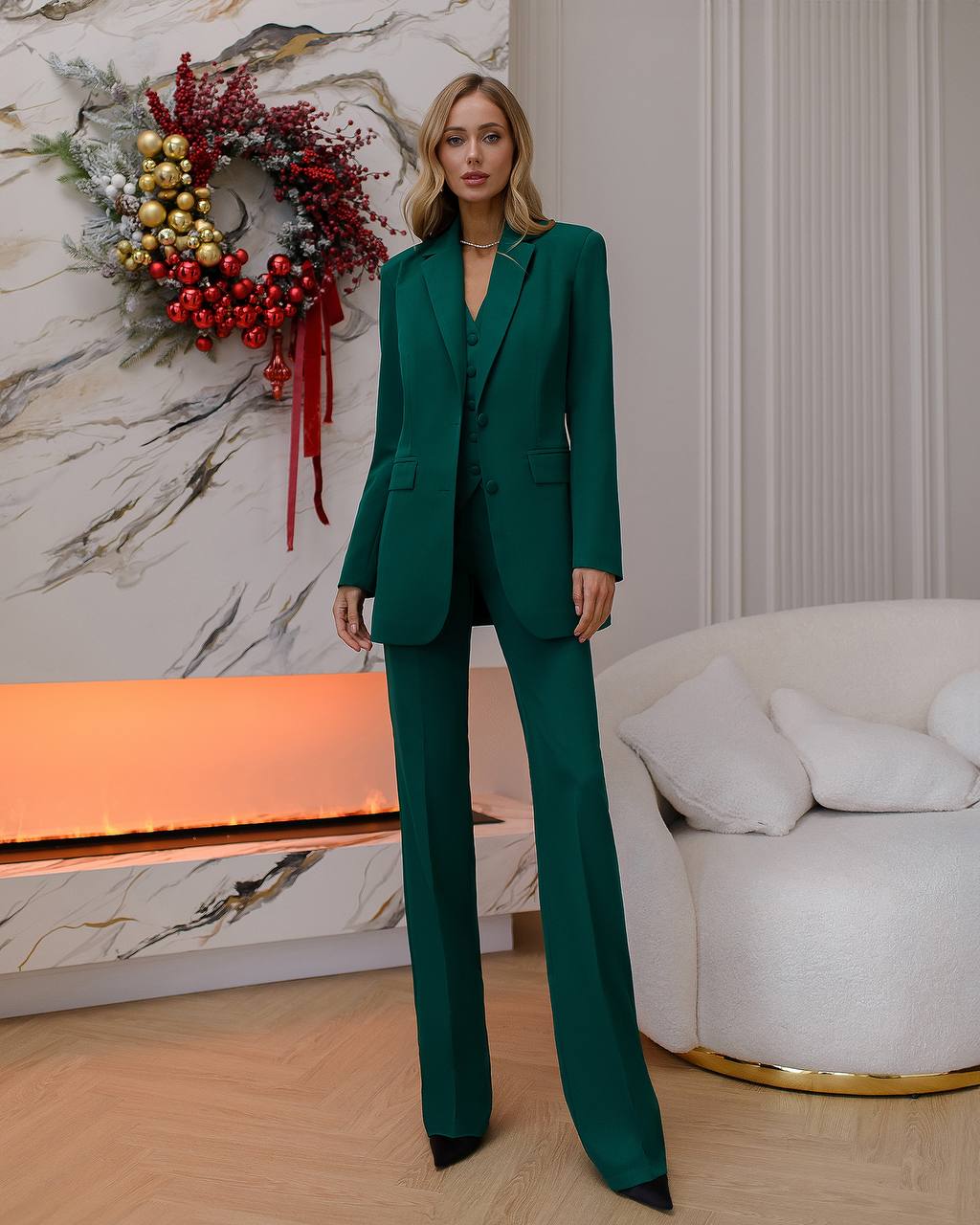 Woman in a green suit standing in a stylish room with a decorative wreath on the wall.