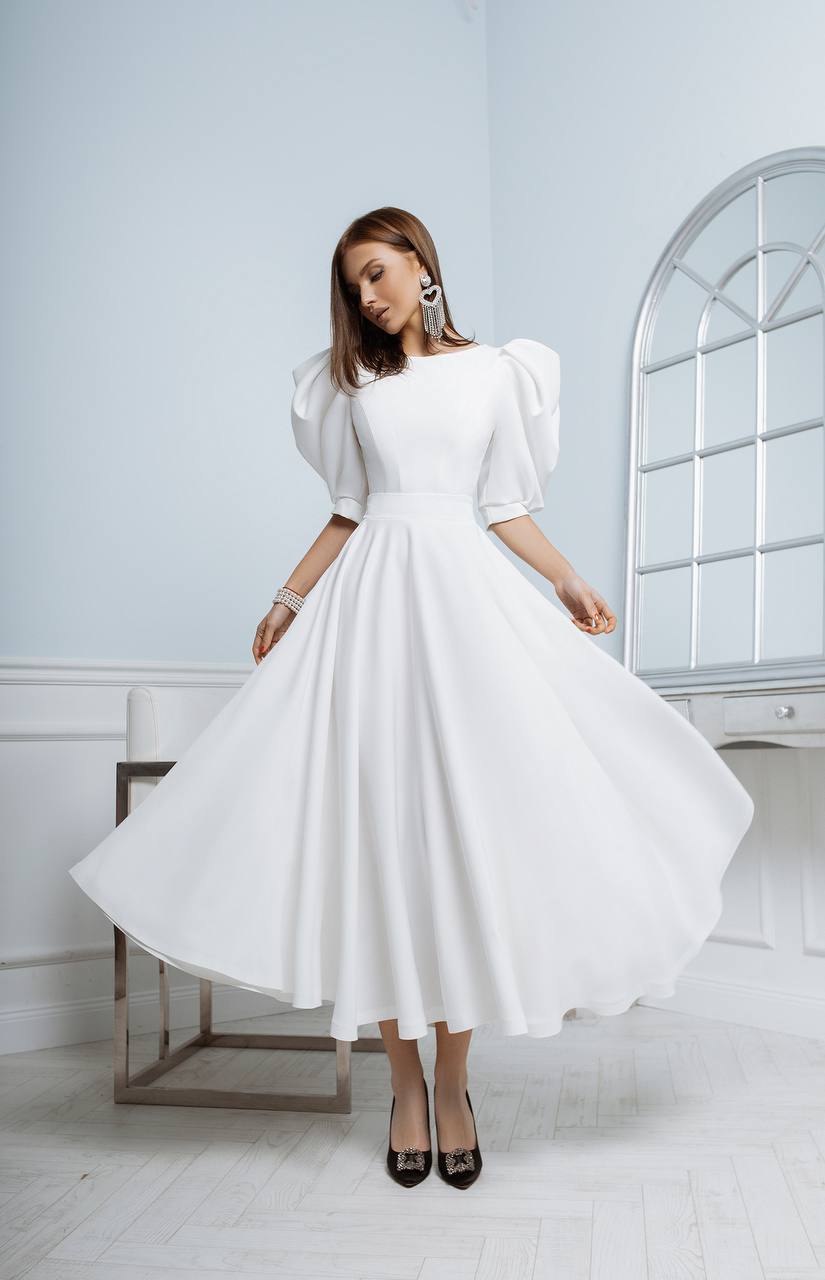 Woman wearing a white dress with puffed sleeves in a minimalistic room.
