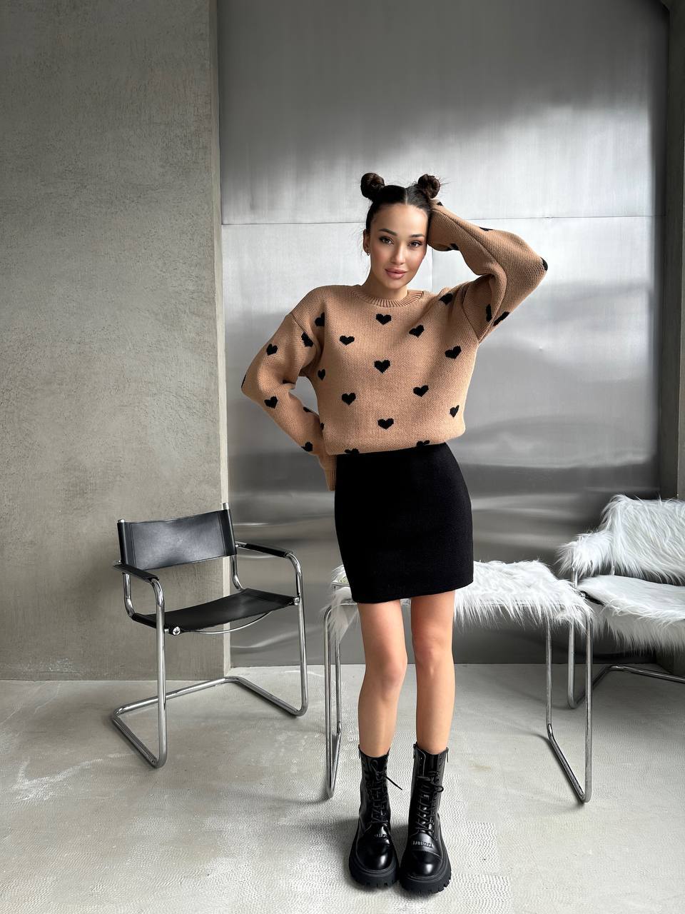 Person wearing a beige sweater with black heart patterns, black skirt, and black boots in a modern room.